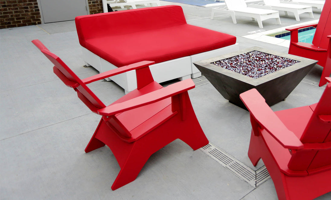 Ledge Loungers red deck chairs and Affinity Square Sunbed with Backrest Cushion surround a square modern fire pit on a concrete poolside patio. The inviting space, complete with white lounge chairs by the pool, is ideal for relaxing.