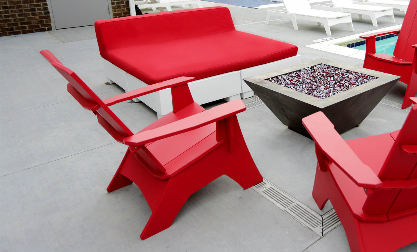Ledge Loungers red deck chairs and Affinity Square Sunbed with Backrest Cushion surround a square modern fire pit on a concrete poolside patio. The inviting space, complete with white lounge chairs by the pool, is ideal for relaxing.