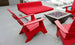 Ledge Loungers red deck chairs and Affinity Square Sunbed with Backrest Cushion surround a square modern fire pit on a concrete poolside patio. The inviting space, complete with white lounge chairs by the pool, is ideal for relaxing.