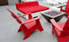 Ledge Loungers red deck chairs and Affinity Square Sunbed with Backrest Cushion surround a square modern fire pit on a concrete poolside patio. The inviting space, complete with white lounge chairs by the pool, is ideal for relaxing.