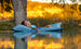 A person with long hair relaxes on a Ledge Lounger Laze Pillow in a serene body of water, surrounded by trees and a golden landscape under warm light, enjoying perfect outdoor comfort.