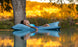 A person with long hair relaxes on a Ledge Lounger Laze Pillow in a serene body of water, surrounded by trees and a golden landscape under warm light, enjoying perfect outdoor comfort.