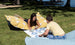 Two kids play a giant lawn game of Ledge Loungers Checkers & Tic Tac Toe on grass, relaxing on bean bags with circular yellow patterns. Trees and water form a serene backdrop under the clear sky.