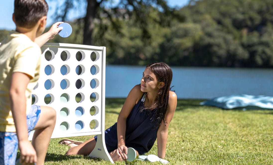 A boy and girl are enjoying oversized outdoor fun with Ledge Loungers Four In A Row by the lake. The boy inserts a disc on the giant board while the girl crouches nearby, observing him. Trees gently sway along the waters edge in the background.