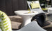 A modern patio setting includes sleek, curved chairs with green patterned cushions and a white pedestal table. Complementing the green theme, an Autograph Tall Ice Bin Side Table by Ledge Lounger ensures refreshments are handy as sunlight casts shadows on the grey tiled floor.
