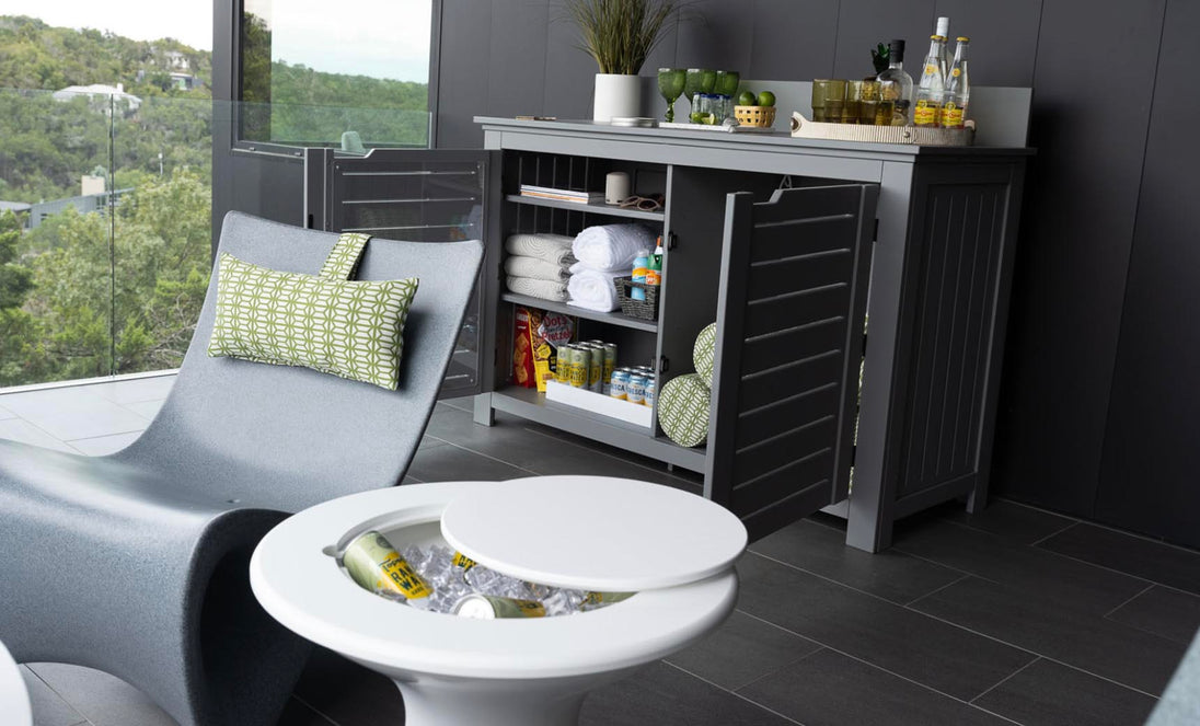 A modern patio showcases a Mainstay Bar Credenza containing towels, glasses, jars, and drinks. Nearby, a sleek gray Ledge Lounger with a green patterned cushion rests beside a round white side table holding a cooler with bottles, set against a scenic outdoor backdrop.