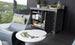 A modern patio showcases a Mainstay Bar Credenza containing towels, glasses, jars, and drinks. Nearby, a sleek gray Ledge Lounger with a green patterned cushion rests beside a round white side table holding a cooler with bottles, set against a scenic outdoor backdrop.