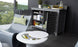A modern patio showcases a Mainstay Bar Credenza containing towels, glasses, jars, and drinks. Nearby, a sleek gray Ledge Lounger with a green patterned cushion rests beside a round white side table holding a cooler with bottles, set against a scenic outdoor backdrop.