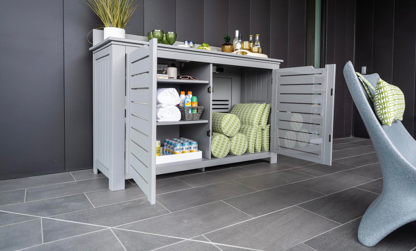 The Mainstay Bar Credenza by Ledge Lounger features open slatted doors revealing towels, beverages, and snacks. Decorative items and plants adorn it, set on a gray tiled floor beside a modern gray chair. Final Sale.
