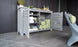 The Mainstay Bar Credenza by Ledge Lounger features open slatted doors revealing towels, beverages, and snacks. Decorative items and plants adorn it, set on a gray tiled floor beside a modern gray chair. Final Sale.