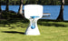 A modern Hoopstr basketball hoop by Ledge Lounger, featuring a blue rim and net, stands on a grassy lawn by the lake. Ideal for a backyard game, trees and a dock accentuate the scene under the clear blue sky.