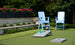 Two light blue Adirondack chairs sit on artificial grass near a Ledge Lounger Cornhole set, with flower beds and green shrubs in the background.