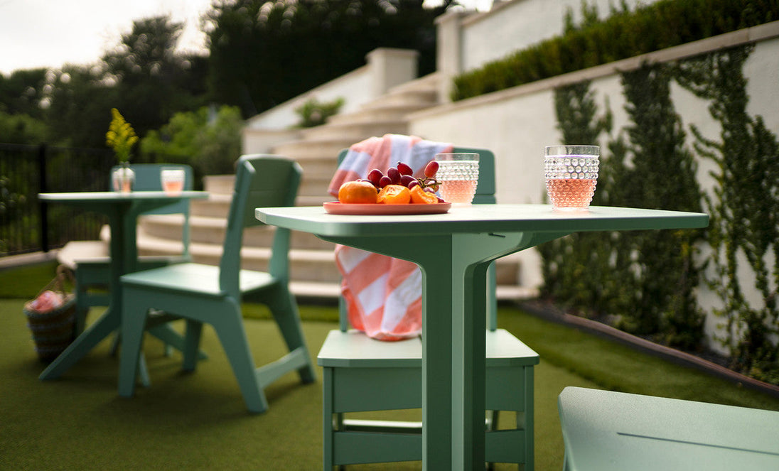 The outdoor space showcases a Mainstay Square Bistro Table by Ledge Lounger with green chairs on the lawn. The table holds a plate of peaches and grapes next to a pink drink in a textured glass, with a striped towel draped over a chair. Lush greenery borders the patio steps.