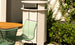 A modern patio scene includes a light green slatted chair beside a round wooden table. A sleek, light outdoor heater is surrounded by lush plants in the background, with sunlight casting gentle shadows nearby.