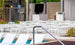 A contemporary outdoor poolside features white Ledge Lounger chairs facing a TV on a stand, surrounded by potted plants, a stainless steel railing, and a dry bar under a roof with wooden decking in the foreground.