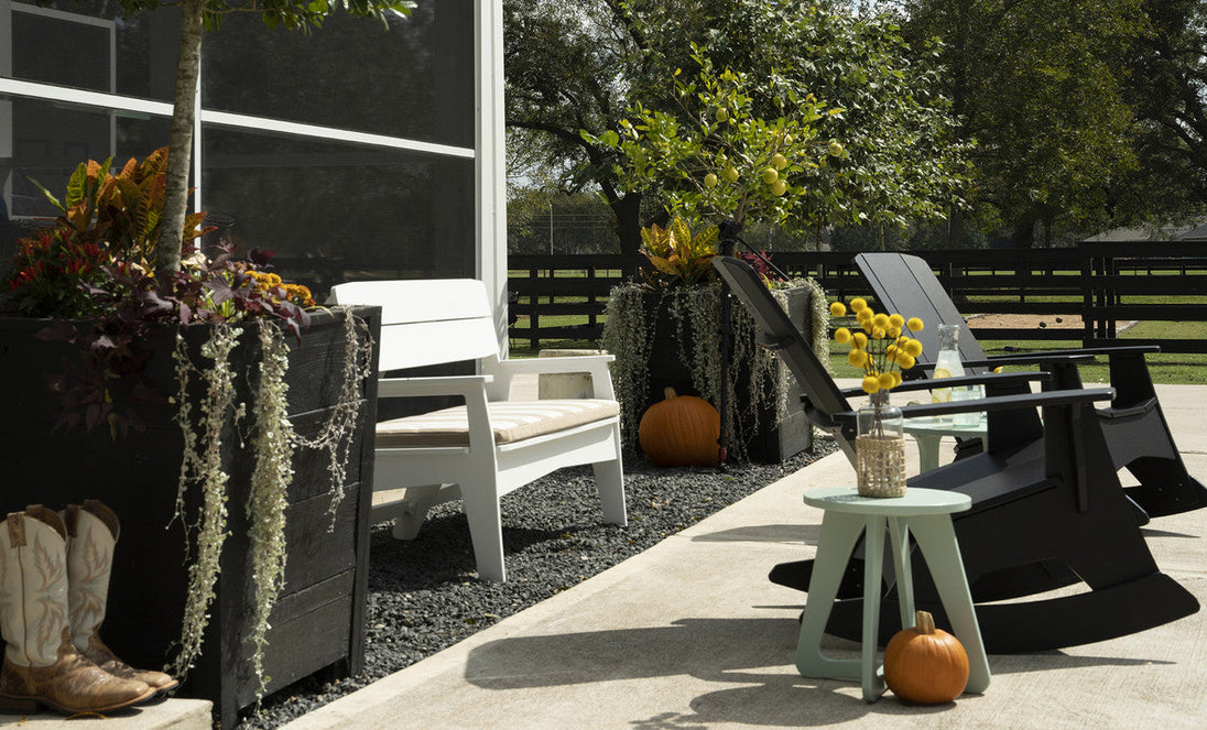 A patio features a white bench with a Mainstay Bench Cushion by Ledge Lounger, dark chairs, and large planters filled with greenery and pumpkins. Cowboy boots rest near the edge, while a fenced grassy area with trees enhances the harmony of the setting.