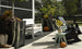 A patio features a white bench with a Mainstay Bench Cushion by Ledge Lounger, dark chairs, and large planters filled with greenery and pumpkins. Cowboy boots rest near the edge, while a fenced grassy area with trees enhances the harmony of the setting.