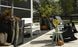 A patio features a white bench with a Mainstay Bench Cushion by Ledge Lounger, dark chairs, and large planters filled with greenery and pumpkins. Cowboy boots rest near the edge, while a fenced grassy area with trees enhances the harmony of the setting.