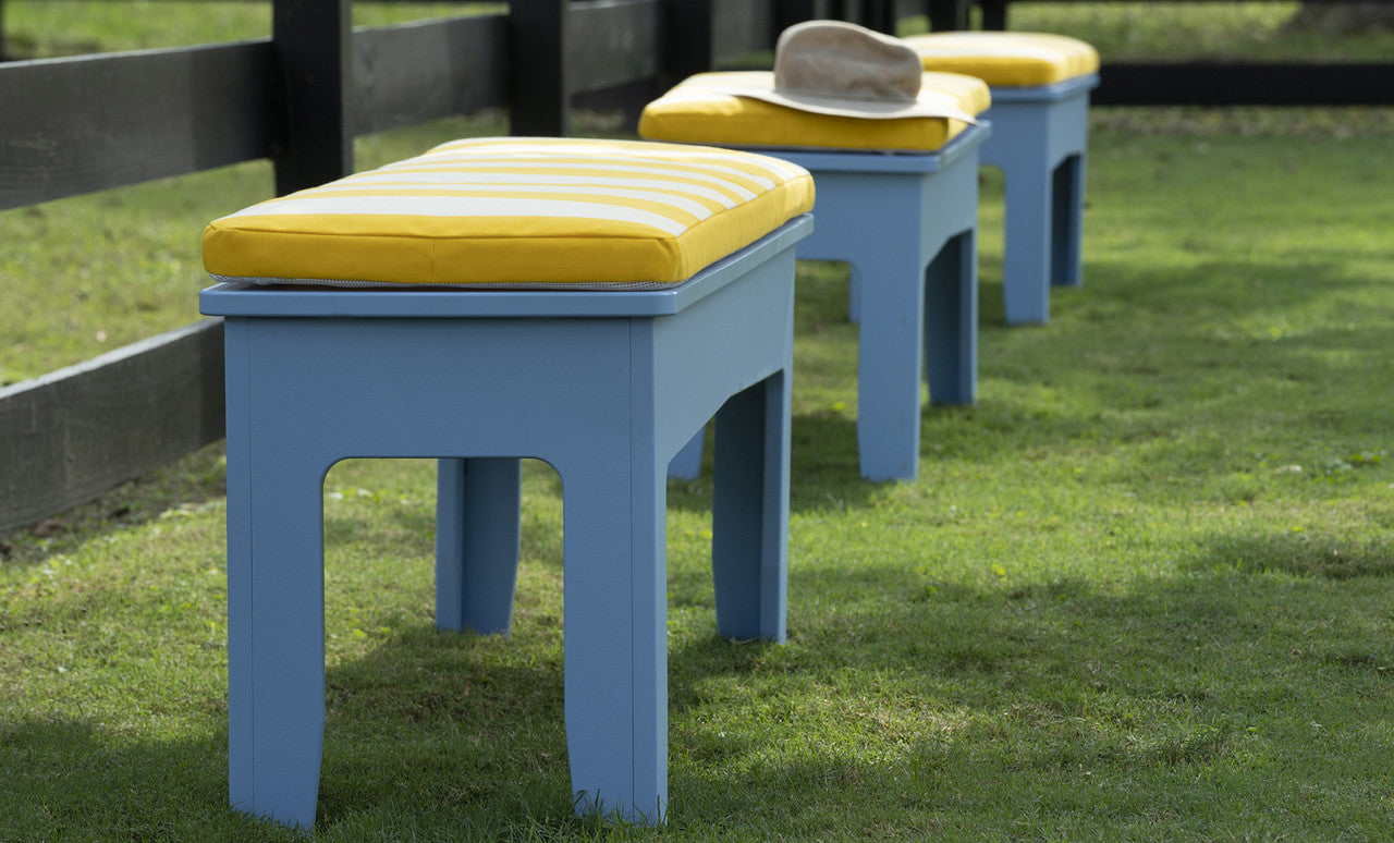 Three Mainstay Dining Benches, 25 by Ledge Lounger, with blue frames and yellow striped cushions are arranged on green grass near a black wooden fence. A tan hat is placed on the bench in front.