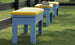 Three Mainstay Dining Benches, 25 by Ledge Lounger, with blue frames and yellow striped cushions are arranged on green grass near a black wooden fence. A tan hat is placed on the bench in front.