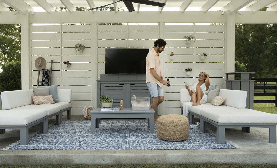 A man and woman relax on outdoor white sofas with a patterned rug; he serves drinks from an ice chest on the table. The area features a white slatted backdrop, wall-mounted TV, and decorative straw hat. Brand products visible: Ledge Lounger (no Mainstay Trash Bin in sight).