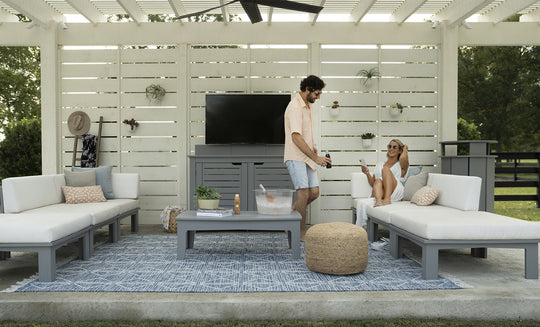 A man and woman relax on outdoor white sofas with a patterned rug; he serves drinks from an ice chest on the table. The area features a white slatted backdrop, wall-mounted TV, and decorative straw hat. Brand products visible: Ledge Lounger (no Mainstay Trash Bin in sight).