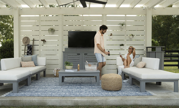 A man and woman relax on outdoor white sofas with a patterned rug; he serves drinks from an ice chest on the table. The area features a white slatted backdrop, wall-mounted TV, and decorative straw hat. Brand products visible: Ledge Lounger (no Mainstay Trash Bin in sight).