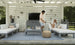 A man and woman relax on outdoor white sofas with a patterned rug; he serves drinks from an ice chest on the table. The area features a white slatted backdrop, wall-mounted TV, and decorative straw hat. Brand products visible: Ledge Lounger (no Mainstay Trash Bin in sight).