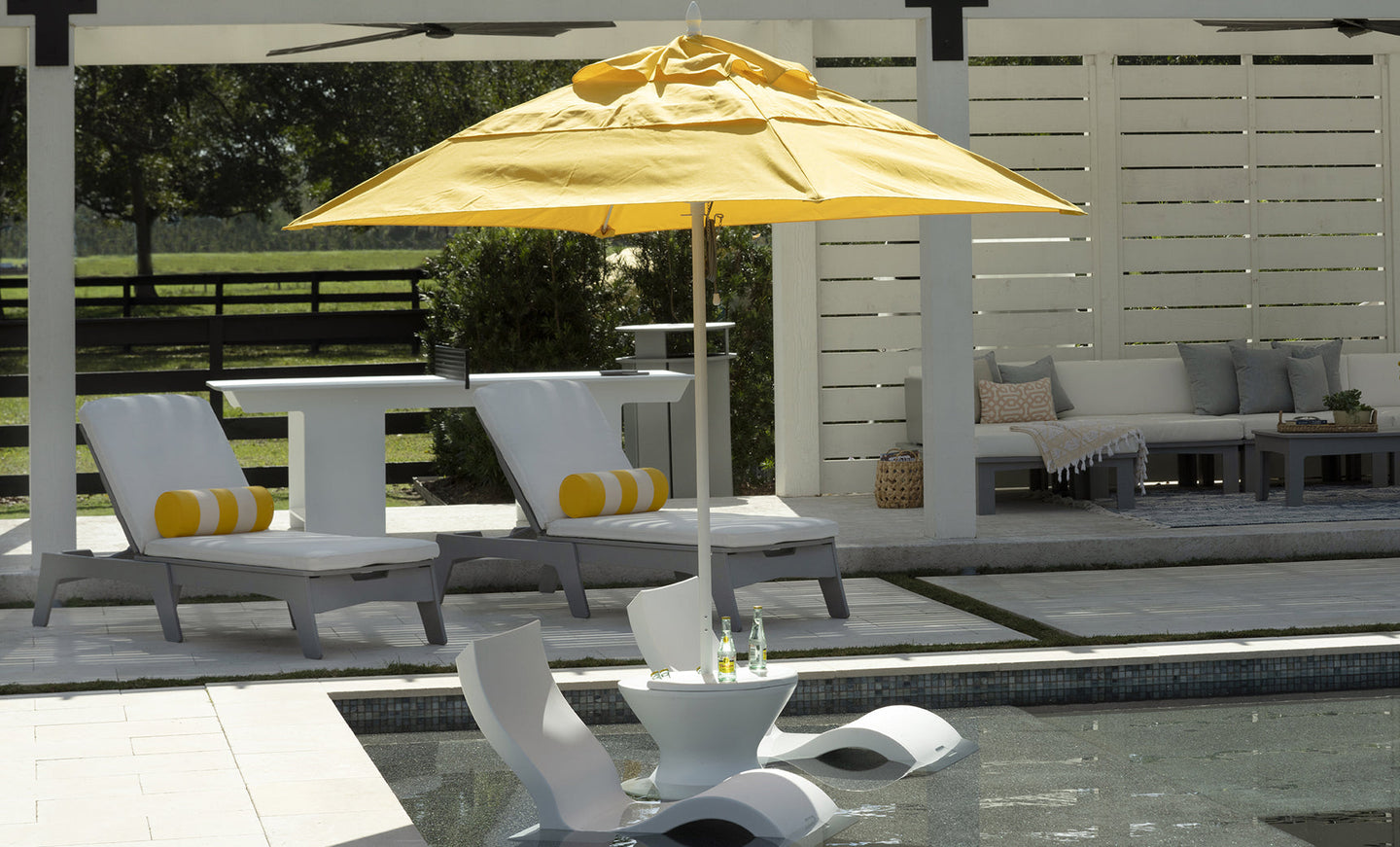Pool scene features Ledge Lounger chairs partially submerged in the water, with a small round table holding drinks under a yellow umbrella. The background includes a seating area with cushions.