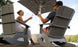 Two people unwind in gray Mainstay Adirondack Rockers by Ledge Lounger on a wooden deck, toasting with bottles under a large umbrella. Theyre near water amid lush greenery, enjoying their sunny and serene outdoor space.