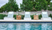 Four Echo Chaise loungers by Ledge Lounger, featuring blue cushions and white towels, are arranged beside a sparkling pool. Wooden side tables are placed between the chairs, set against a backdrop of lush green trees and shrubs for a serene outdoor ambiance.