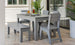The setup includes a gray patio table with matching chairs and the Mainstay Dining Bench 25 from Ledge Lounger. The table features tableware, a plant centerpiece, and a glass bottle. Large windows and potted plants complete the serene environment.