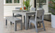 The setup includes a gray patio table with matching chairs and the Mainstay Dining Bench 25 from Ledge Lounger. The table features tableware, a plant centerpiece, and a glass bottle. Large windows and potted plants complete the serene environment.