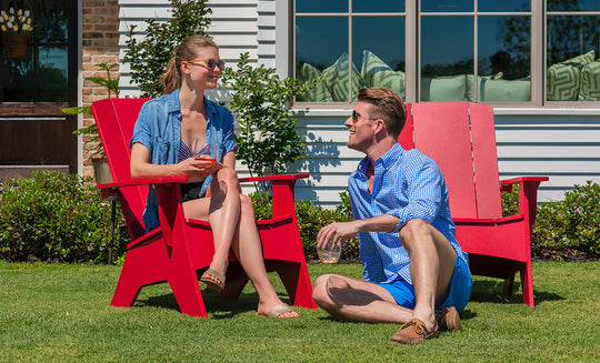 A woman sits on a Mainstay Ultimate Adirondack from Ledge Lounger, drink in hand, while a man relaxes nearby. Both don sunglasses and casual summer wear, enjoying the sunny garden bliss with chic outdoor lounge chairs in front of their houses large windows.