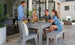 Four people unwind on a patio by a light-colored house. Two sit on the Mainstay Dining Bench 25 from Ledge Lounger, while the others stand, chatting and enjoying drinks near glass doors and potted plants.