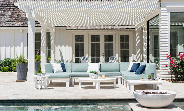 Under a white pergola, an outdoor patio features a light blue L-shaped sofa in high-quality fabric and a white round table. Nearby, a large flower planter enhances the scene. A fire pit adds warmth to the serene pool atmosphere, complemented by Ledge Loungers Mainstay Adirondack Headrest Pillow.