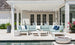 Under a white pergola, an outdoor patio features a light blue L-shaped sofa in high-quality fabric and a white round table. Nearby, a large flower planter enhances the scene. A fire pit adds warmth to the serene pool atmosphere, complemented by Ledge Loungers Mainstay Adirondack Headrest Pillow.