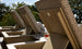 Several Echo Chaise loungers from Ledge Lounger, featuring geometric-patterned cushions, are placed in a serene outdoor space. A glass of brown liquid rests on one lounger’s arm. Tall trees rise in the background under a clear sky.