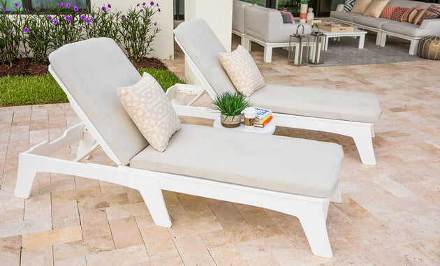 Two Mainstay Chaise Cushions by Ledge Lounger provide full-body comfort on a tiled patio in white outdoor lounge chairs with beige cushions and patterned pillows. A small round table holds a plant and books amid purple flowers and lantern seating, offering a tranquil ambiance for relaxation.