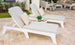 Two Mainstay Chaise Cushions by Ledge Lounger provide full-body comfort on a tiled patio in white outdoor lounge chairs with beige cushions and patterned pillows. A small round table holds a plant and books amid purple flowers and lantern seating, offering a tranquil ambiance for relaxation.