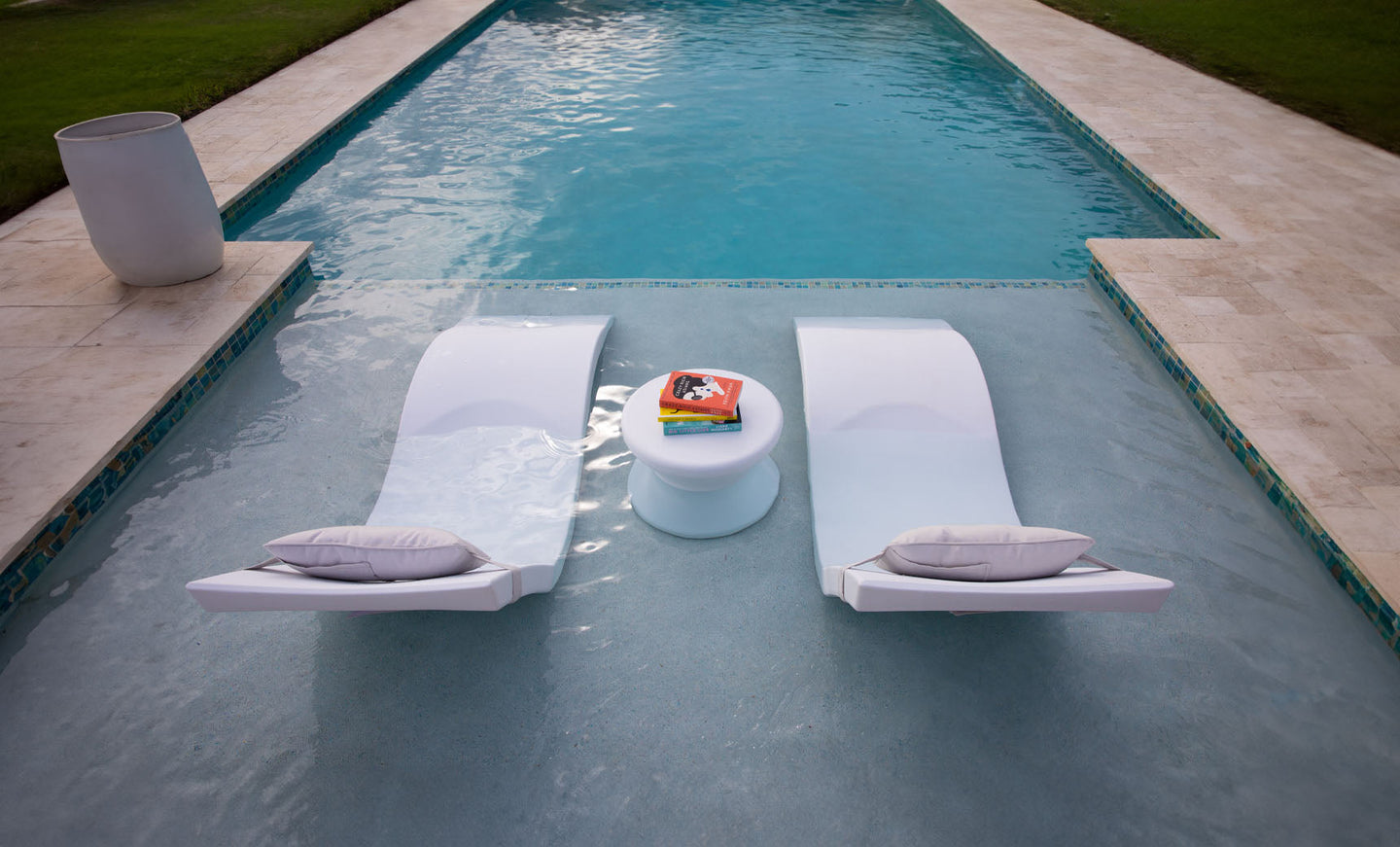 Two Signature In-Pool Chaise Loungers with cushions from Ledge Lounger are partially submerged in a pool, flanking a small table with magazines. This Outdoor Paradise In-Pool Furniture Bundle is set amidst beige stone decking and lush green grass.