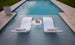 Two Signature In-Pool Chaise Loungers with cushions from Ledge Lounger are partially submerged in a pool, flanking a small table with magazines. This Outdoor Paradise In-Pool Furniture Bundle is set amidst beige stone decking and lush green grass.