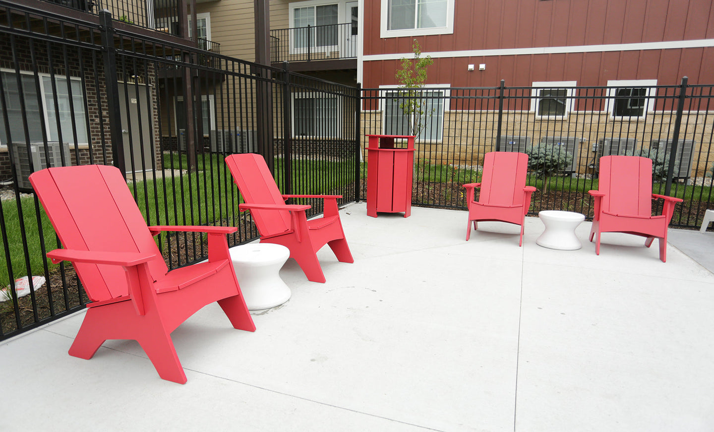 Four red Adirondack chairs form a semi-circle on a concrete patio, flanked by two white side tables. The space is bordered by a black metal fence, with apartment buildings and balconies in view.