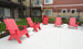 Four red Adirondack chairs form a semi-circle on a concrete patio, flanked by two white side tables. The space is bordered by a black metal fence, with apartment buildings and balconies in view.