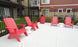 Four red Adirondack chairs form a semi-circle on a concrete patio, flanked by two white side tables. The space is bordered by a black metal fence, with apartment buildings and balconies in view.