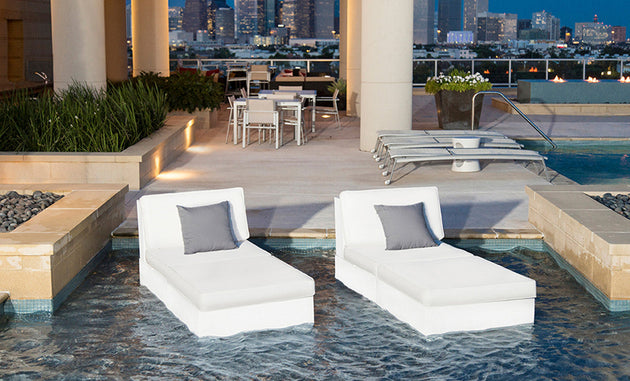 Two white Ledge Lounger chairs with gray Throw Pillow 18 Square - Final Sale pillows float in a modern rooftop pool. Nearby, theres a patio table and chairs on the deck with the city skyline visible in the background.