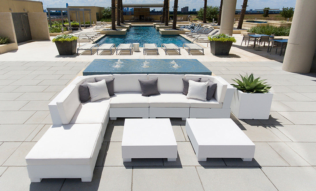 On a modern patio, white sectional sofas with Ledge Lounger Throw Pillow 18 Square (Final Sale) in gray, face a blue pool. Two white square coffee tables and a large planter with succulents sit on the gray tile floor. Lounge chairs are nearby.
