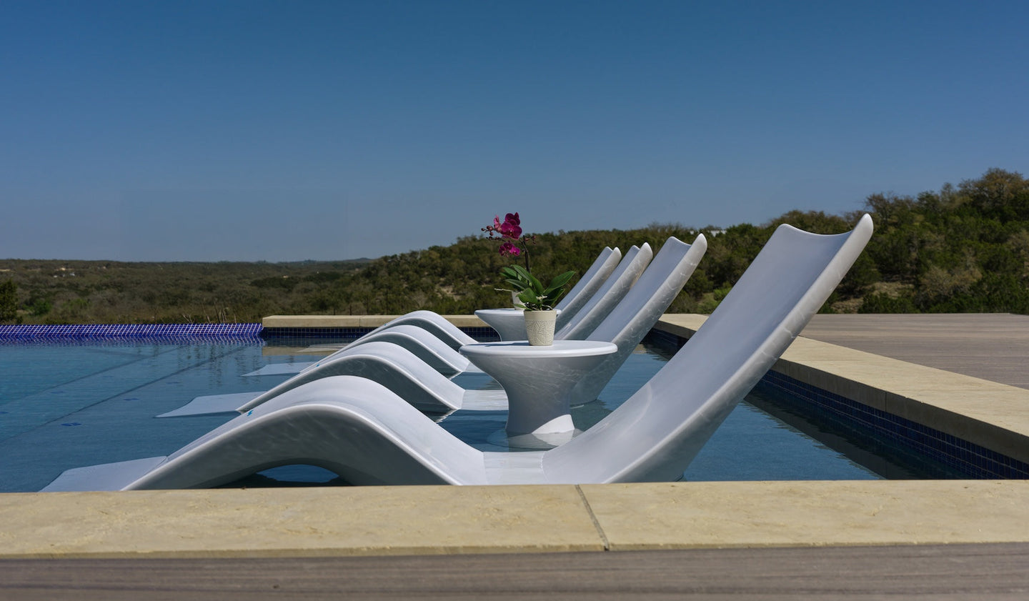 Four white lounge chairs are partially submerged in a pool beside the Ledge Lounger Autograph Tall Ice Bin Side Table with a potted purple orchid, overlooking green hills under a clear blue sky.
