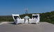 Two modern white chairs with black-and-white striped cushions sit on a wooden deck. Between them rests the Ledge Lounger Autograph Tall Ice Bin Side Table, topped with a potted orchid and books. Green trees and blue sky complete the serene scene.