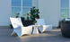 Two modern white chairs and a Ledge Lounger Autograph Standard Side Table with a potted orchid are arranged on a wooden patio deck, surrounded by green plants and large windows in the background.